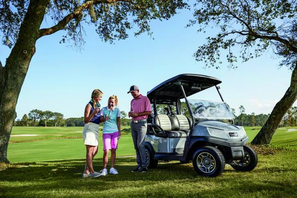 Three golfers stand in front of a golf buggy on a golf course and discuss the game.